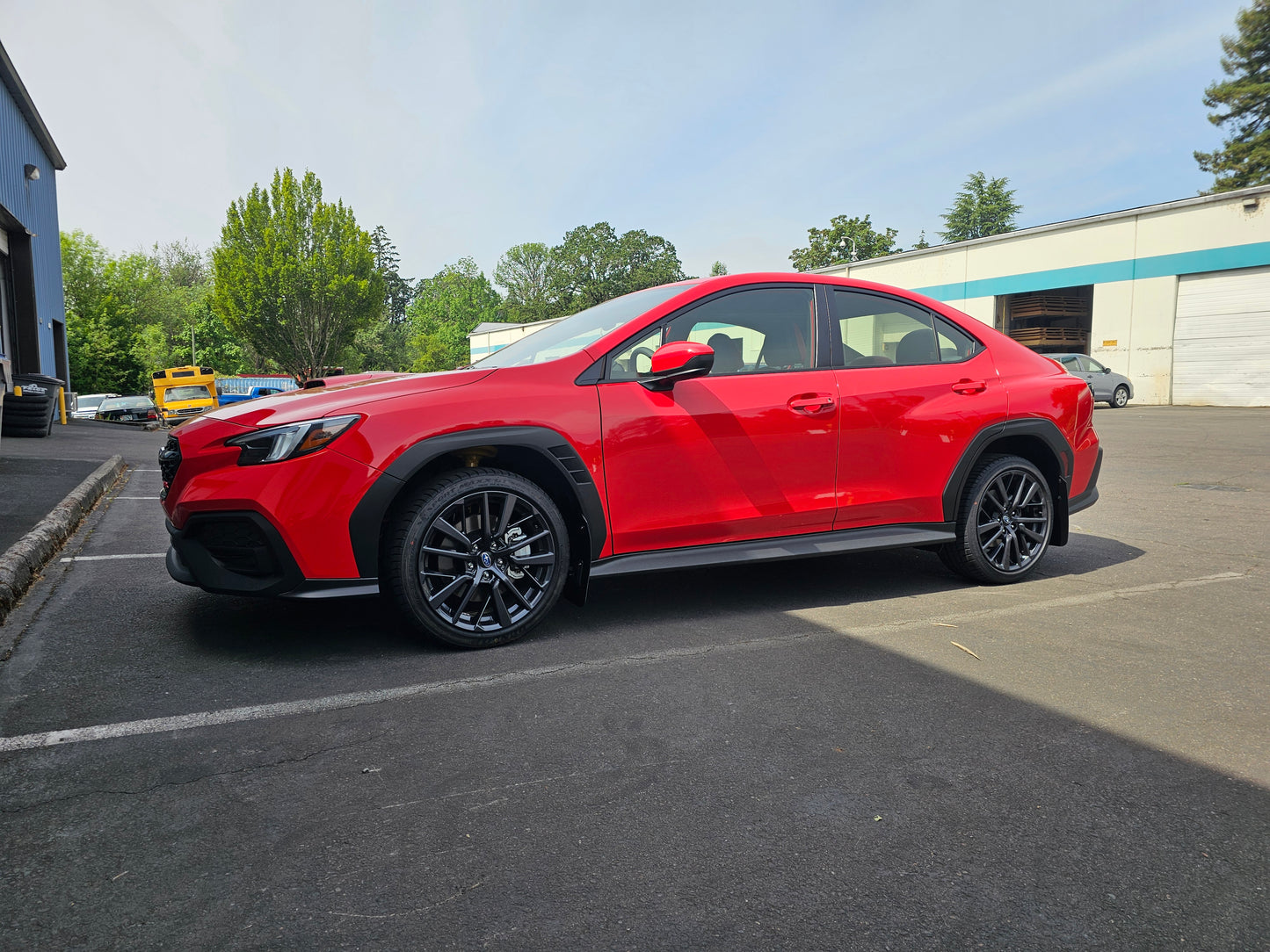 A red 1.25" lifted WRX parked in an outdoor lot. The vehicle features black multi-spoke wheels, black lower trim, and black B-pill caps, photographed against a background of a light blue sky, trees, and industrial buildings.