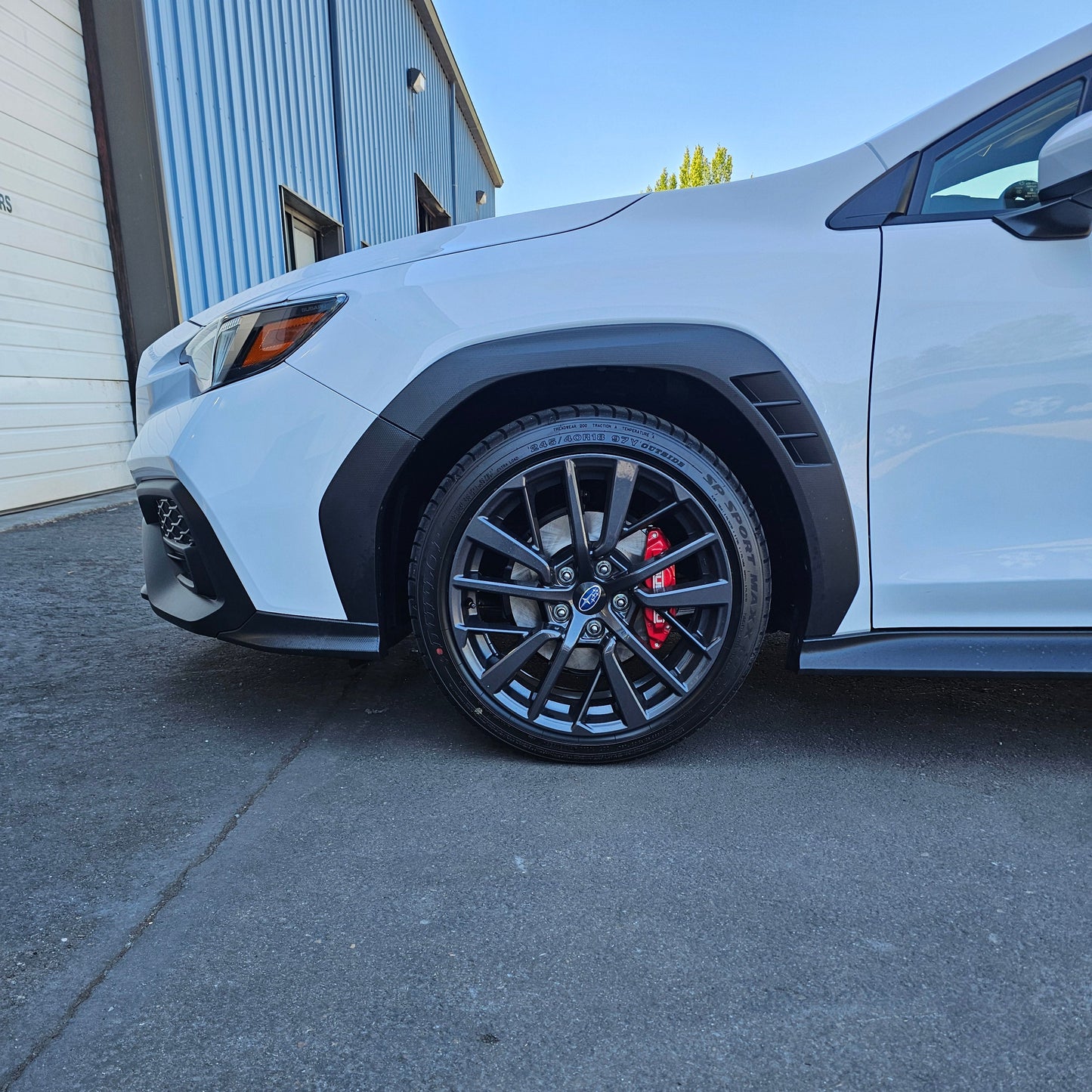 A close-up side view of a white WRX lifted 1.25" front wheel and fender, featuring black multi-spoke alloy wheels with red brake calipers, parked on asphalt next to a blue and white building.