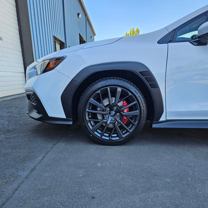 A close-up side view of a white WRX lifted 1.25" front wheel and fender, featuring black multi-spoke alloy wheels with red brake calipers, parked on asphalt next to a blue and white building.