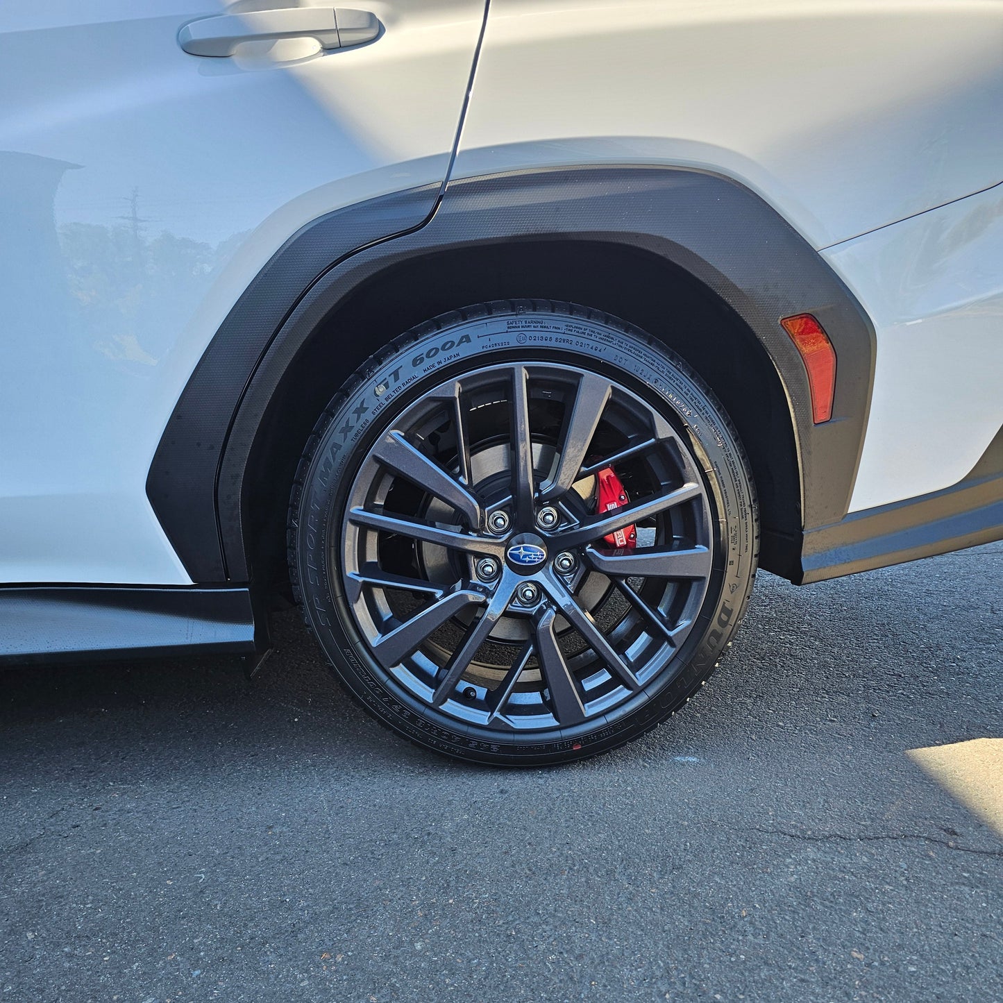 Close-up of a white WRX's 1.25" lifted front wheel featuring a dark gray multi-spoke alloy tire, black wheel arch, and red brake calipers, parked on asphalt.