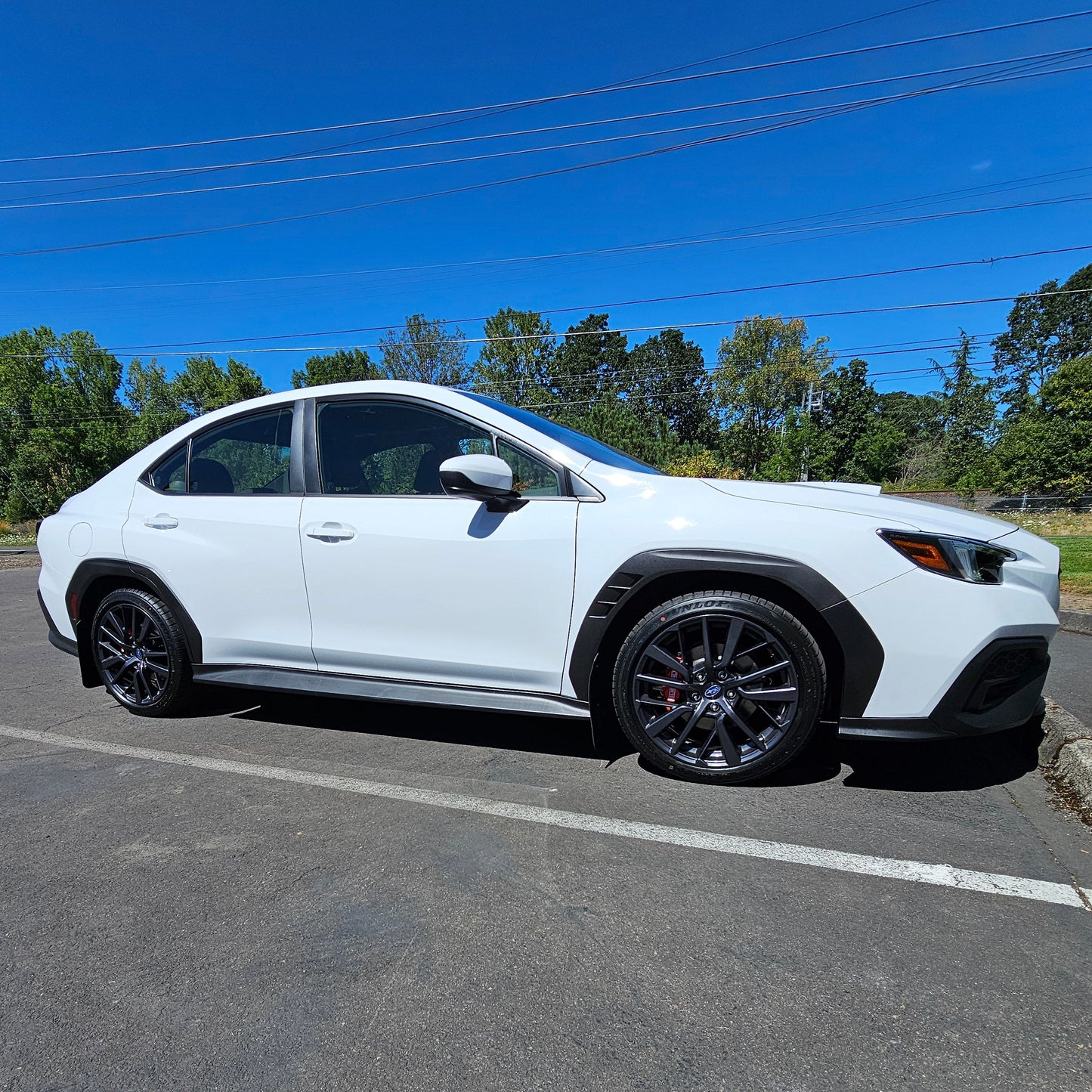 A white WRX Lifted 1.25 Inches parked on an asphalt lot under a clear blue sky, with trees and utility poles in the background.